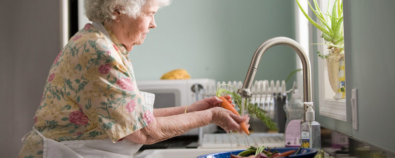 ältere Damen mit Schürze steht an der Spüle und wäscht Gemüse